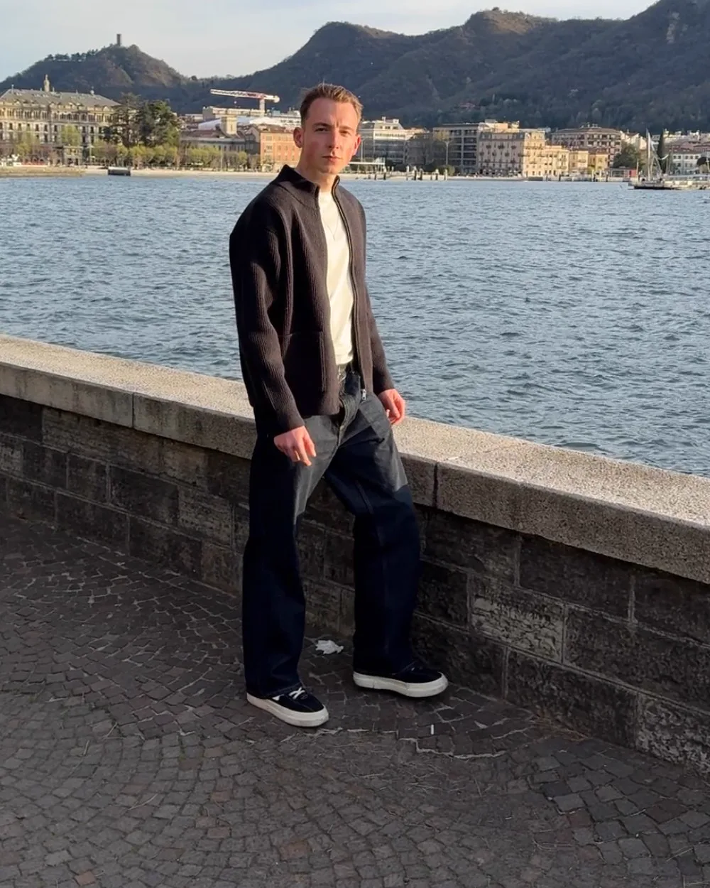Robin Heinicke an der Uferpromenade des Comer Sees, in Strickjacke, entspannt an eine Mauer gelehnt